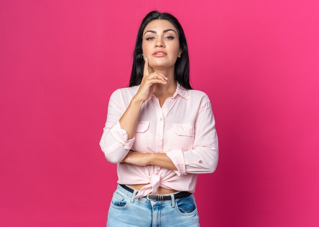 Mujer hermosa joven en ropa casual con expresión seria de confianza con una sonrisa en la cara inteligente de pie sobre la pared rosa