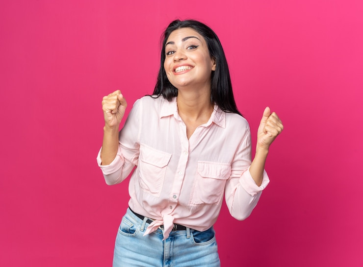 Foto gratuito mujer hermosa joven en ropa casual feliz y emocionado apretando los puños de pie en rosa