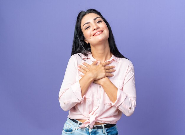 Mujer hermosa joven en ropa casual feliz y positiva tomados de la mano en el pecho sintiéndose agradecido de pie sobre la pared azul