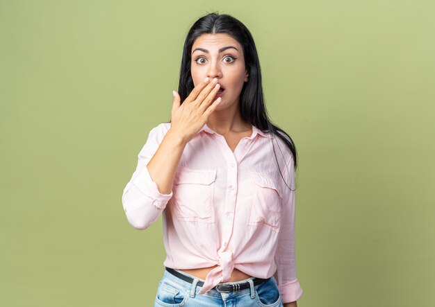 Mujer hermosa joven en ropa casual mirando al frente conmocionado cubriendo la boca con la mano de pie sobre la pared verde