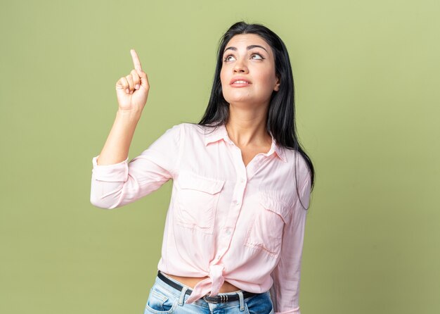 Mujer hermosa joven en ropa casual mirando hacia arriba apuntando con el dedo índice a algo sonriente de pie sobre la pared verde