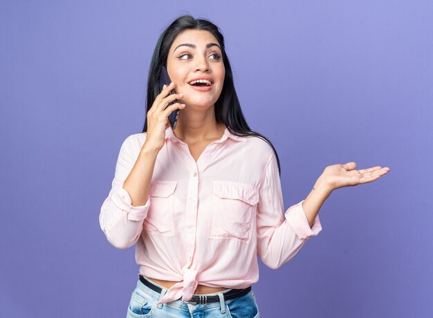 Mujer hermosa joven en ropa casual sonriendo alegremente feliz y positivo mientras habla por teléfono móvil de pie sobre la pared azul