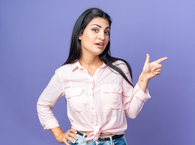 Mujer hermosa joven en ropa casual sonriendo confiado apuntando con el dedo índice hacia el lado de pie sobre la pared azul