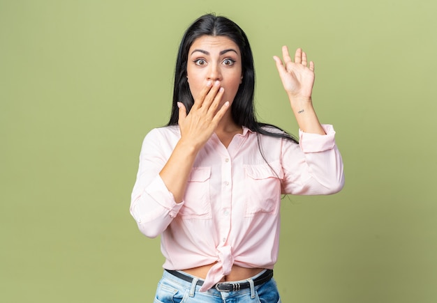 Mujer hermosa joven en ropa casual sorprendida y conmocionada cubriendo la boca con la mano de pie sobre la pared verde