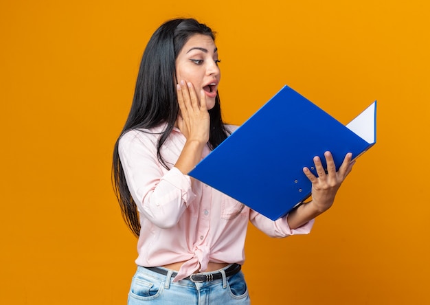 Mujer hermosa joven en ropa casual sosteniendo la carpeta mirándola feliz y sorprendida de pie en naranja