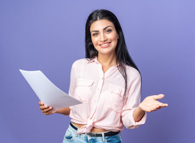 Mujer hermosa joven en ropa casual sosteniendo documentos haciendo gesto de bienvenida con la mano sonriendo de pie amistoso sobre la pared azul