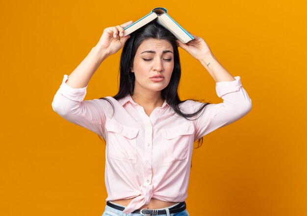 Mujer hermosa joven en ropa casual sosteniendo el libro sobre su cabeza está molesto de pie en naranja