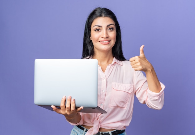 Mujer hermosa joven en ropa casual sosteniendo portátil feliz y positivo mirando sonriendo confiado mostrando pulgar hacia arriba