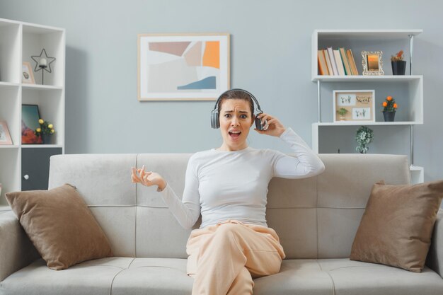 Mujer hermosa joven con ropa informal sentada en un sofá con auriculares mirando a la cámara confundida y disgustada levantando el brazo con disgusto tratando de quitarse los auriculares para escuchar algo