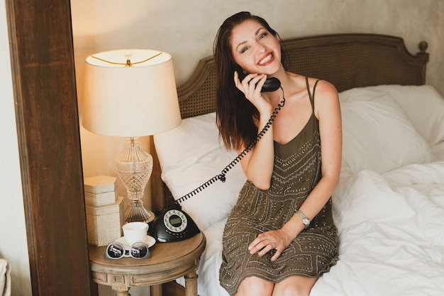 Mujer hermosa joven sentada en la cama en el hotel, elegante vestido de noche, estado de ánimo sensual, hablando por teléfono, sonriendo, coqueta, mirando, sexy