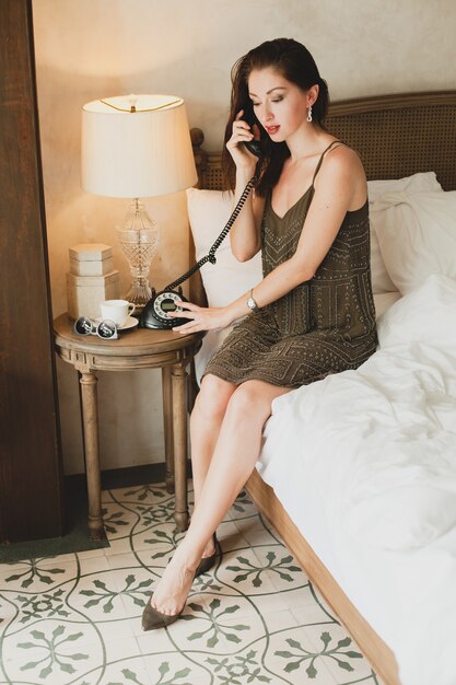 Mujer hermosa joven sentada en la cama en el hotel, elegante vestido de noche, estado de ánimo sensual, hablando por teléfono, sonriendo, coqueta, mirando, sexy