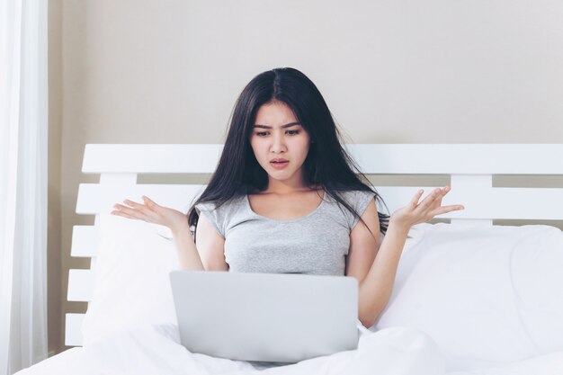 Mujer hermosa joven sentada en la cama, usa una computadora portátil y se siente seria y triste