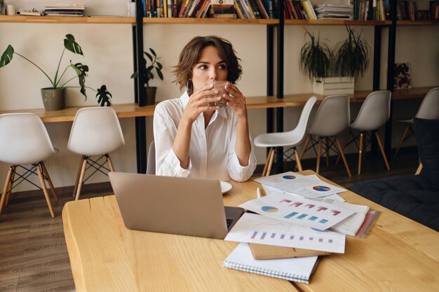 Mujer hermosa joven sentada en la mesa con una computadora portátil y diagramas mientras toma café en la oficina moderna