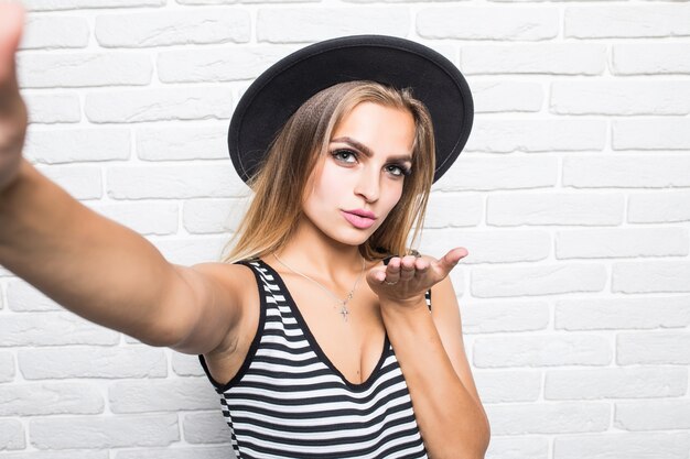 Mujer hermosa joven con sombrero de paja beso de golpe mientras toma selfie en teléfono aislado en la pared de ladrillo