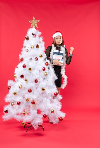 Mujer hermosa joven con sombrero de santa claus y de pie detrás del árbol de Navidad decorado sosteniendo regalos y mirando