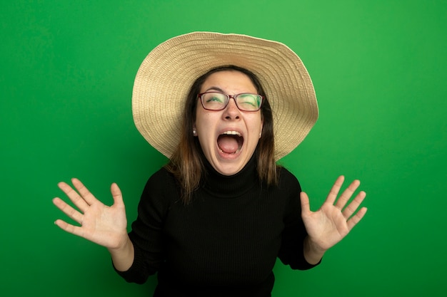 Mujer hermosa joven con sombrero de verano en un cuello alto negro y gafas gritando con las manos levantadas de pie sobre la pared verde