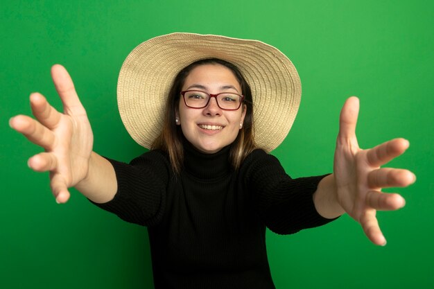 Mujer hermosa joven con sombrero de verano en un cuello alto negro y gafas haciendo gesto de bienvenida feliz y positivo sonriendo amistoso de pie sobre la pared verde