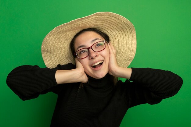 Mujer hermosa joven con sombrero de verano en un cuello alto negro y gafas mirando hacia arriba feliz y positivo sonriendo alegremente de pie sobre la pared verde