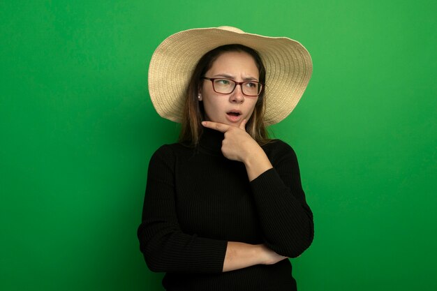 Mujer hermosa joven con sombrero de verano en un cuello alto negro y gafas mirando a un lado con la mano en la barbilla confundida de pie sobre la pared verde