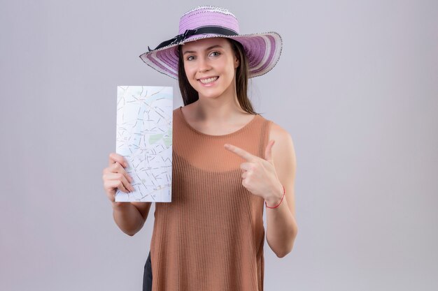 Mujer hermosa joven en sombrero de verano con mapa apuntando hacia ella sonriendo con cara feliz sobre pared blanca