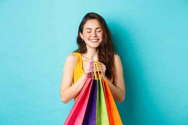 Mujer hermosa joven soñando despierto con ropa nueva, sosteniendo bolsas de la compra, cerrar los ojos y sonreír soñadora, de pie sobre fondo azul.