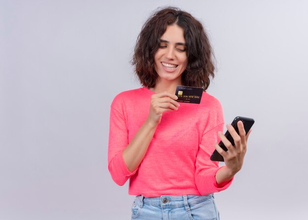 Mujer hermosa joven sonriente que sostiene la tarjeta y el teléfono móvil en la pared blanca aislada con el espacio de la copia