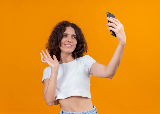 Mujer hermosa joven sonriente que sostiene el teléfono móvil y que lo saluda en la pared anaranjada aislada con el espacio de la copia