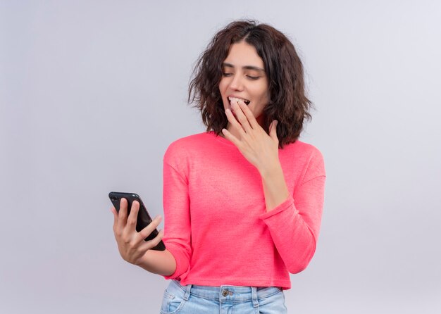 Mujer hermosa joven sonriente que sostiene el teléfono móvil y que pone la mano en el labio en la pared blanca aislada con el espacio de la copia