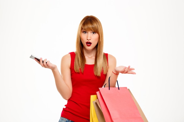 Mujer hermosa joven sorprendida que se coloca con las compras sobre la pared blanca