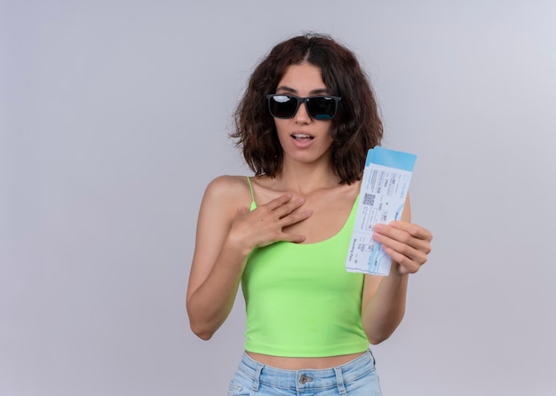 Mujer hermosa joven sorprendida que lleva gafas de sol y que sostiene los billetes de avión en la pared blanca aislada con el espacio de la copia