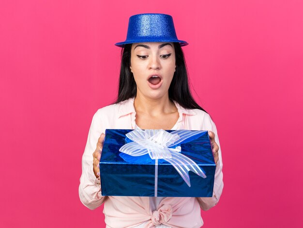 Mujer hermosa joven sorprendida que lleva el sombrero del partido que sostiene y que mira la caja de regalo aislada en la pared rosada