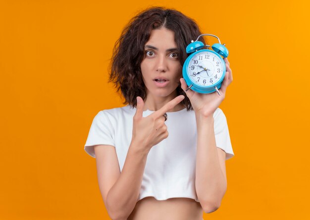 Mujer hermosa joven sorprendida que sostiene el reloj de alarma y que señala en la pared anaranjada aislada con el espacio de la copia