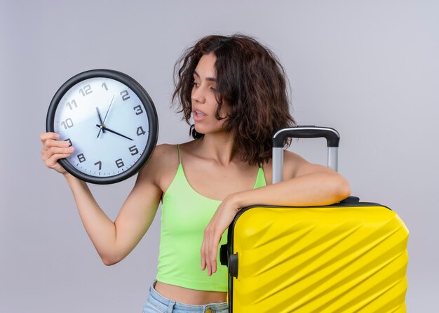 Mujer hermosa joven sorprendida que sostiene el reloj y la maleta del viajero y que mira el reloj en la pared blanca aislada