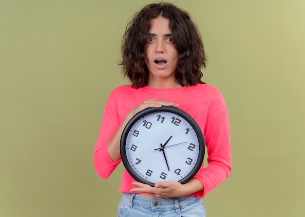 Foto gratuita mujer hermosa joven sorprendida que sostiene el reloj en la pared verde aislada con el espacio de la copia