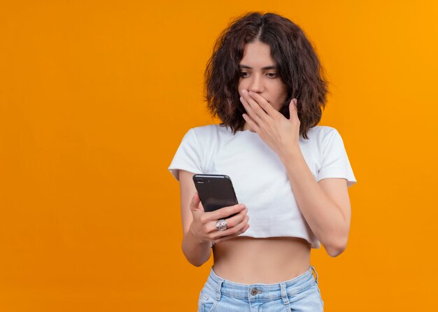 Mujer hermosa joven sorprendida que sostiene el teléfono móvil y que pone la mano en la boca en la pared anaranjada aislada con el espacio de la copia