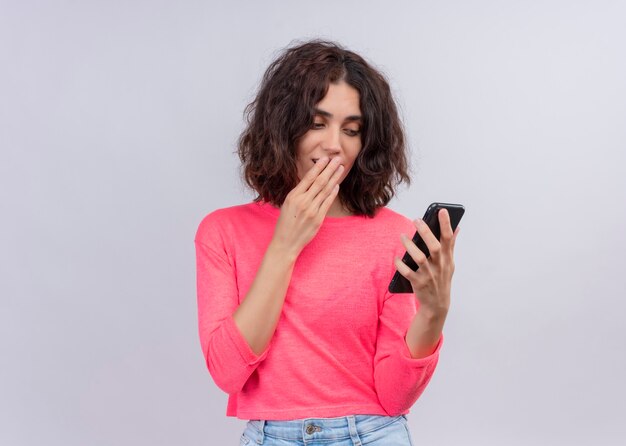 Mujer hermosa joven sorprendida que sostiene el teléfono móvil y que pone la mano en la boca en la pared blanca aislada con el espacio de la copia