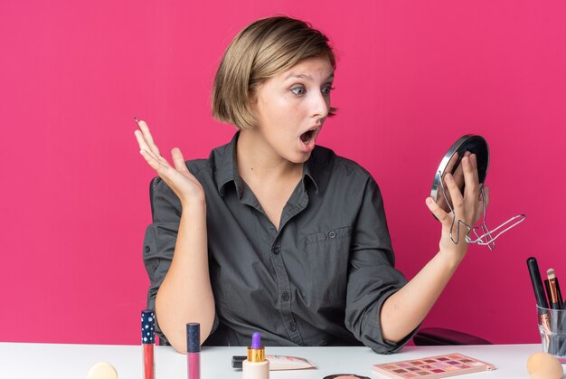 Mujer hermosa joven sorprendida se sienta a la mesa con herramientas de maquillaje sosteniendo pincel de maquillaje mirando el espejo en su mano extendiendo la mano