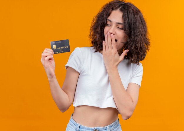 Mujer hermosa joven sorprendida sosteniendo la tarjeta y poniendo la mano en la boca en la pared naranja aislada con espacio de copia