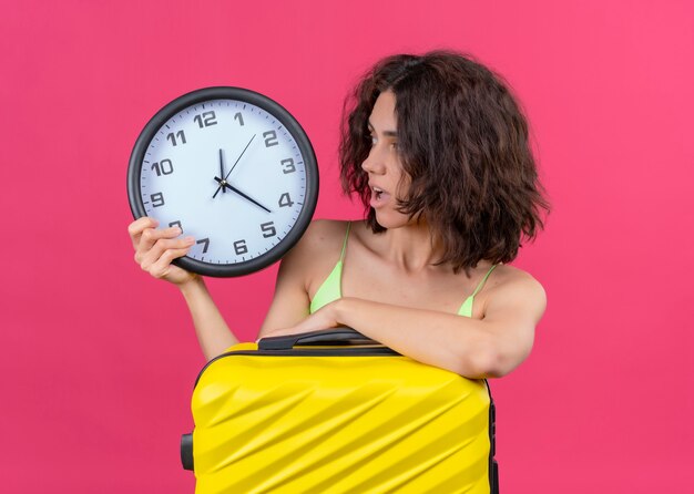 Mujer hermosa joven sorprendida del viajero que sostiene la maleta y el reloj y que mira el reloj en la pared rosada aislada