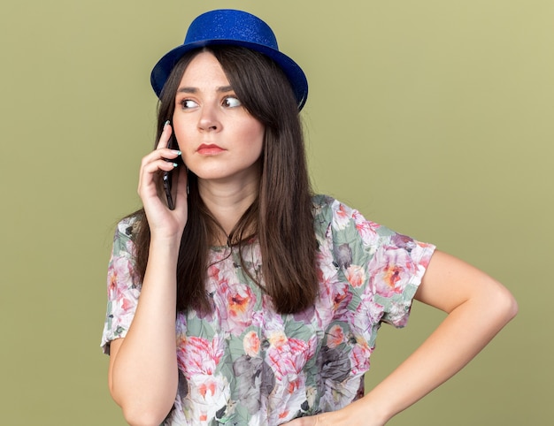Mujer hermosa joven sospechosa que lleva el sombrero del partido que habla en el teléfono que pone la mano en la cadera aislada en la pared verde oliva
