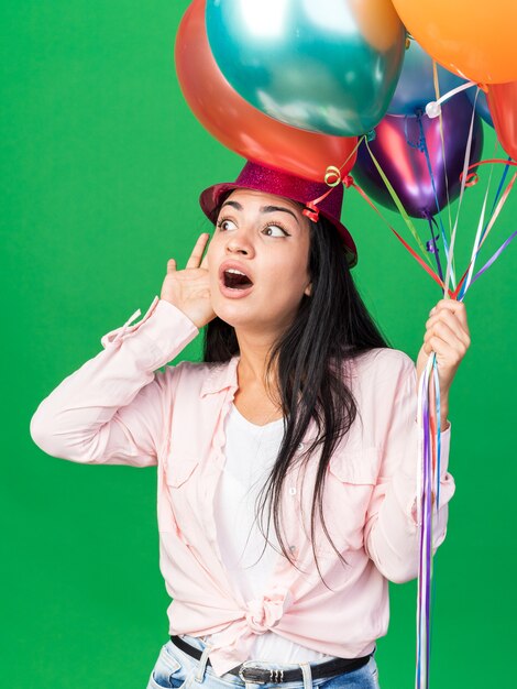 Mujer hermosa joven sospechosa con sombrero de fiesta sosteniendo globos mostrando gesto de escucha aislado en la pared verde