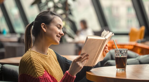 Mujer hermosa joven en suéter anaranjado que lee el libro interesante en café