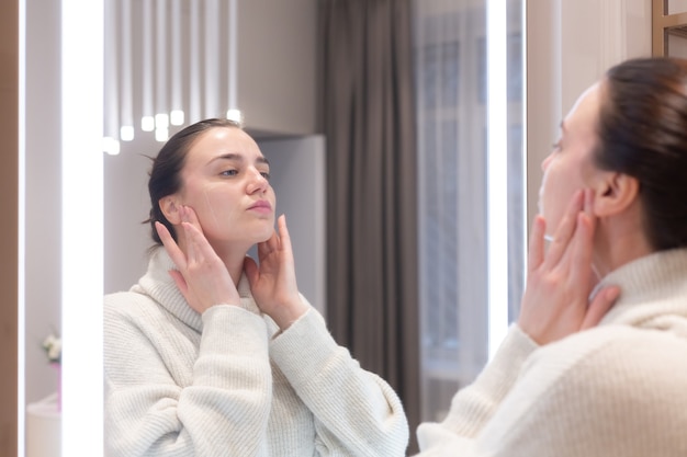 Mujer hermosa joven con un suéter en un salón de belleza se mira en el espejo, se toca la cara, piensa en los próximos procedimientos, se considera a sí misma