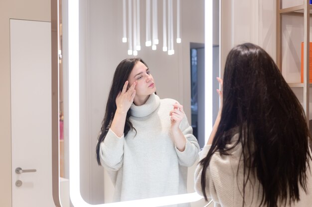 Mujer hermosa joven con un suéter en un salón de belleza se mira en el espejo, se toca la cara, piensa en los próximos procedimientos, se considera a sí misma
