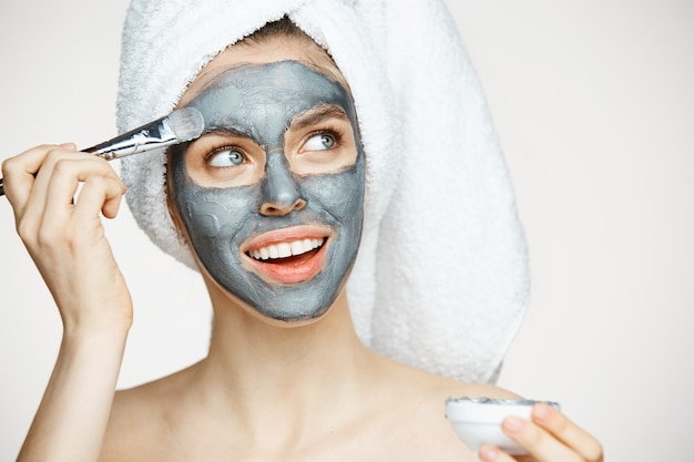 Mujer hermosa joven en toalla en la cabeza que cubre la cara con la máscara sonriendo. Cosmetología de belleza y spa.