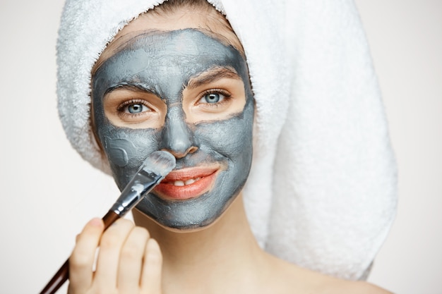 Mujer hermosa joven en toalla en la cabeza que cubre la cara con la máscara sonriendo. Cosmetología de belleza y spa.