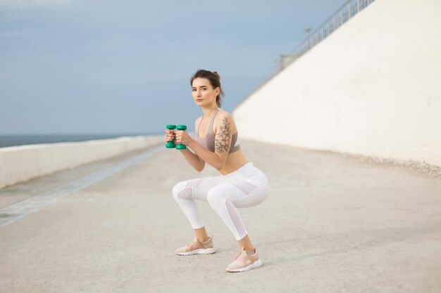 Mujer hermosa joven en top deportivo y polainas blancas haciendo sentadillas con pesas en las manos mientras mira cuidadosamente en cámara al aire libre