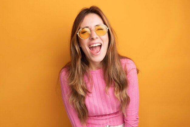 Mujer hermosa joven en top rosa con gafas feliz y emocionado riendo parado sobre naranja