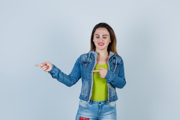 Mujer hermosa joven en traje de mezclilla apuntando hacia la izquierda, cerrando los ojos y mirando alegre, vista frontal.