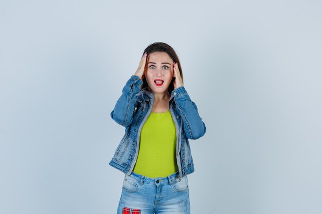 Mujer hermosa joven en traje de mezclilla con las manos en la cabeza y mirando sorprendido, vista frontal.